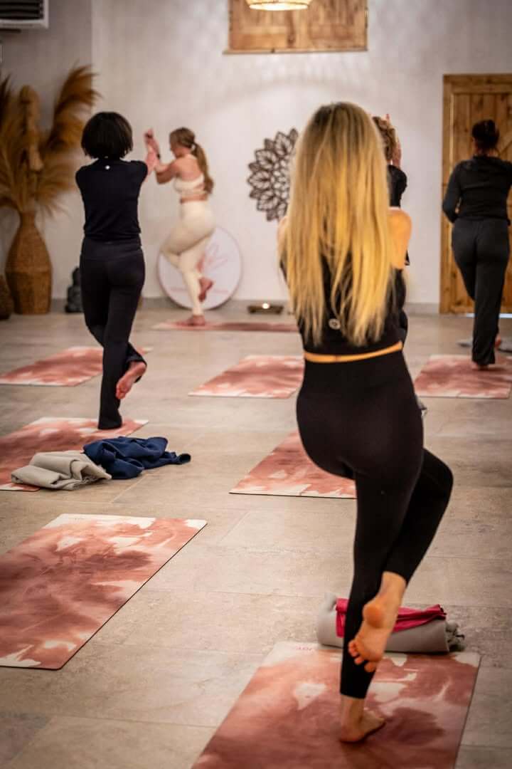 Grupa kobiet praktykujących jogę w pozycji drzewa (vrkshasana) na różowo-białych matach w jasnej sali warsztatowej Masuria Lakehouse z logo Ahimsa Yoga by Iwona w tle