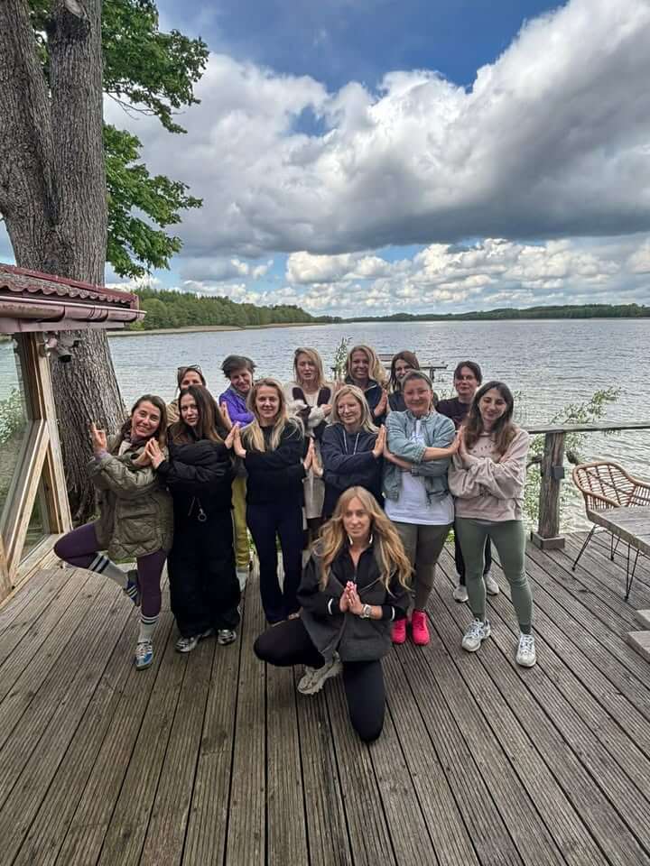 Pamiątkowe zdjęcie grupy uczestniczek na drewnianym pomoście nad mazurskim jeziorem – kobiety stoją z dłońmi złożonymi w geście namaste, prowadząca z przodu w pozycji półklęku, w tle tafla wody i zachmurzone niebo